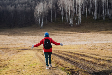Hiker in red jacket arms outstretched embracing landscape, centered stance on muddy track with birch trees framing background, practicing mindful breathing, joy and freedom, wellness travelの写真素材