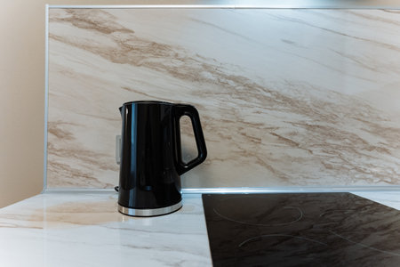 black electric kettle on marble countertop near induction stove, sleek modern appliance with glossy finish and ergonomic handle, morning beverage setup, bright neutral backsplash and subtleの写真素材
