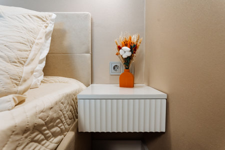 cozy bedside corner with orange vase and soft bed linens, nightstand styled for evening reading and calm mood, ceramic vase holding dried floral arrangement beside outlet and textured headboard.の写真素材