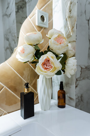 marble vanity with peach bouquet, glass vase and perfume bottles, soft warm lighting from sculpted mirror, white ceramic sink and gold tile accents creating intimate luxury atmosphere for bathroomの写真素材