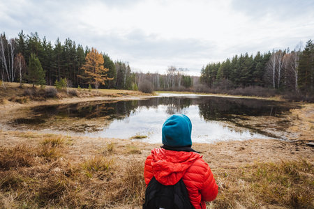 Child observes tranquil nature, Child in bright clothing contemplates serene forest scene by water,...