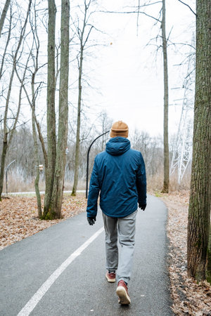 male walking through leafless woods on curving asphalt lane, blue jacket and tan beanie, scouting...