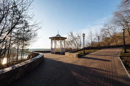 Open Waterfront Gazebo With Paved Promenade. White Pavilion Overlooking Calm Water, Lamp Posts And...