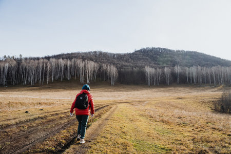 White hiker walking toward distant hills across expansive plain, lone adventurer with backpack and beanie, panoramic scene of birch line, muddy tracks and open sky suggesting long journeyの写真素材