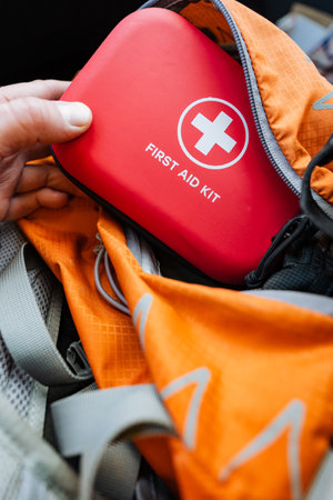 Packing Red First Aid Kit Into Orange Backpack Beside Travel Mug, Outdoor Camping Preparation Showing Hand Stowing Compact Medical Supply With Secure Straps, Pockets And Ready Access For Campsiteの写真素材