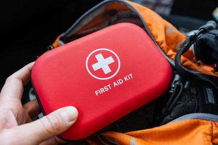 Hand Pulling Red First Aid Kit From Backpack On Trail, Outdoor Hiker Retrieving Compact Medical Kit...