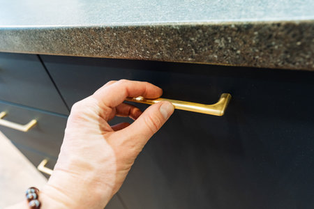 CloseUp Hand On Brass Cabinet Handle, Dark Blue Cabinet Under Stone Countertop, Wristwatch Visible, Morning Light, Homeowner Opening Drawer, Tactile Grip, Clean Contemporary Kitchen Sceneの写真素材