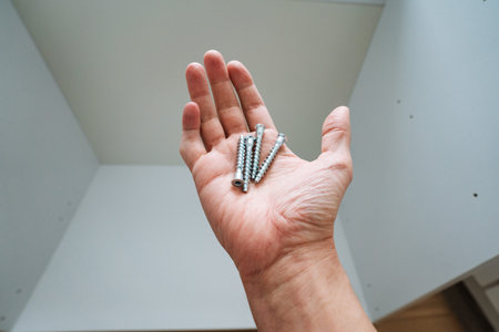 Hand Inspecting Screws For Assembly, DIYer Comparing Fastener Sizes And Threads Under Bright Light Before Fitting, Careful Quality Check With Blurred Furniture Parts In Background.の写真素材