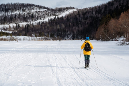 Skier In Yellow Jacket Heading Toward Tree Line Across Wide Snow Corridor, Layered Hills And Forest Edges, Steady Pace And Thoughtful Mood, Overcast Sky And Crisp Air During Winter Ski Trek.の写真素材