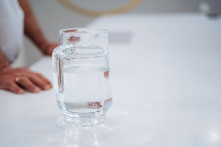 Glass Pitcher Filled With Water Hands Resting On White Countertop, Caregiver Preparing Simple Refreshment For Client In Bright Kitchen, Clear Glass Catching Soft Natural Light, Calm Minimalistの写真素材