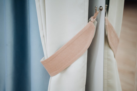 Closeup Curtain Tieback Holding Drapes, Beige Fabric Strap Fastened To Metal Hook Between White And Blue Panels, Soft Natural Light Casting Gentle Shadow, Visible Stitching And Texture, Modernの写真素材
