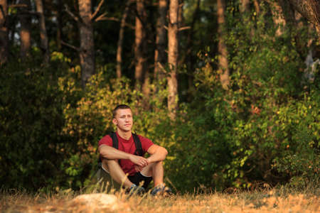 Happy hiker man is resting, sitting on the ground and looking forward, against a background of pine...