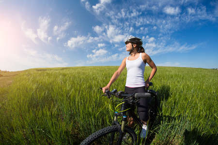 Cheerful beautiful young woman in white shirt with a bike and helmet on a field smiling against blue skyの写真素材