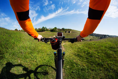 Hands in orange jacket holding handlebar of a bicycle with green meadow on backgroundの写真素材