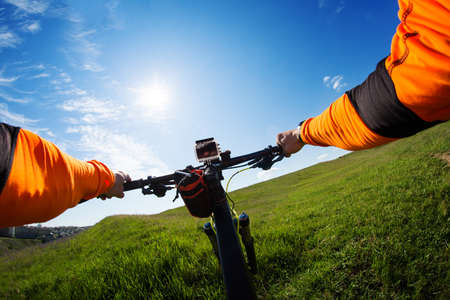 Hands in orange jacket holding handlebar of a bicycle with green meadow on background