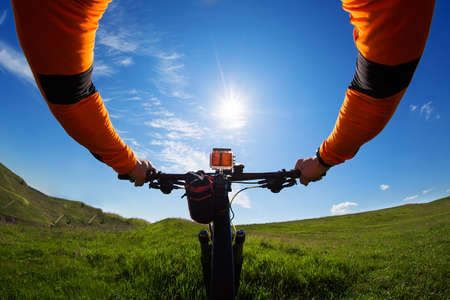 Hands in orange jacket holding handlebar of a bicycle with green meadow on backgroundの写真素材