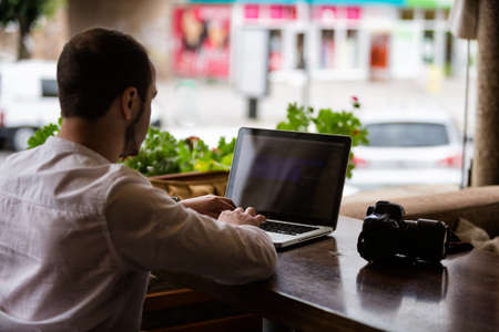 Young photographer in casual clothes with camera sitting in caffee open air and working with laptop