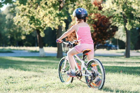 Little brunette girl in helmet riding bicycle in the park at sunset. back viewの写真素材