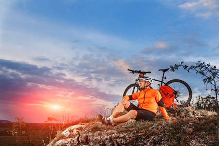 Biker resting on top of mountain with his bicycle near him. Cyclist riding outdoor on sunset. Healthy lifestyle concept.の写真素材