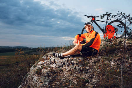 Biker resting on top of mountain with his bicycle near him. Cyclist riding outdoor on sunset. Healthy lifestyle concept.の写真素材