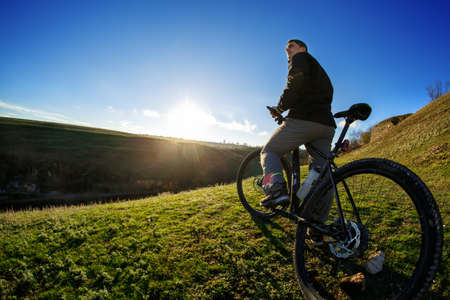 low angle view of cyclist standing with mountain bike at sunrise against bright sun and blue sky. sping season. Horisontal wide angle fisheye photoの写真素材