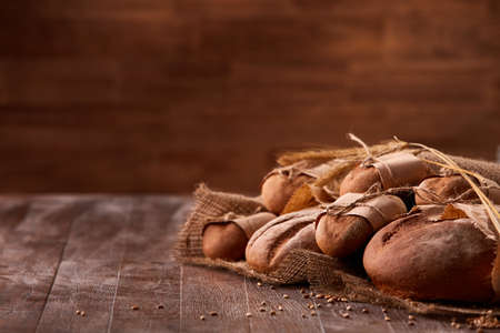 different bread on the wooden table, paper bags, burlap, rope and ears of wheat with brown blurred...