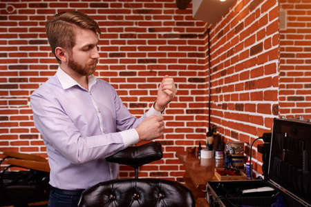 Portrait of the hairdresser. Cheerful young bearded man fastens the buttons on the sleeve on his shirt.の写真素材