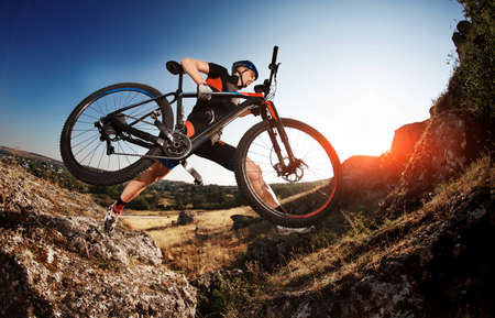 Professional Cyclist Taking his Bike up the Rocky Trail at evening. Extreme Sport Concept. Low angle...