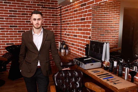 Portrait of handsome young hairdresser standing at barber shop near armchair, hairstylist puts one hand on the chair.の写真素材