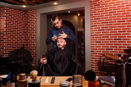 Young handsome barber combing attractive man while sitting at the chair and looking at phone in barbershop.の写真素材