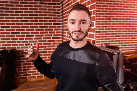 Handsome bearded hairdresser in stylish black wear is holding a scissors while standing at the barbershop.の写真素材