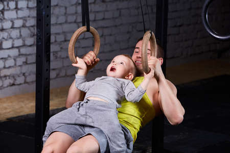 Happy father and little son exercising with gimnastic rings and smiling against brick wall in the...