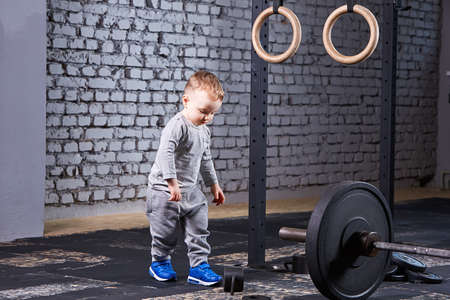 Little cute boy in the grey sportwera standing near barbell against brick wall in the cross fit gym.の写真素材