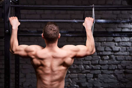 Rear view photo of muscular male doing exercises on horizontal bar against brick wall at the cross...