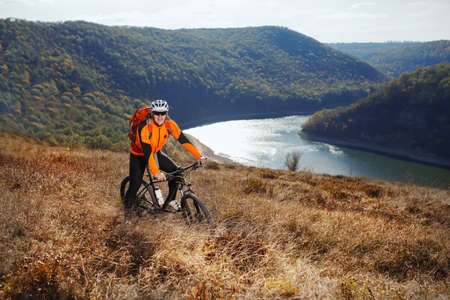 Cyclist in orange jacketr stands with his bike under river against beautiful landscape with mountain.の写真素材