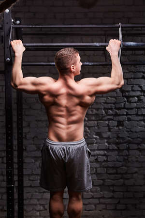 Rear view photo of the young muscular male doing exercises on horizontal bar against brick wall at...