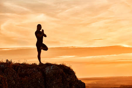 Silhouette of the man practicing yoga on the hill against beautiful sunset. Attractive man in the sportwear. Summer or spring season in the countryside. Horizontal photo. Concept of the healthy lifestyle.の写真素材