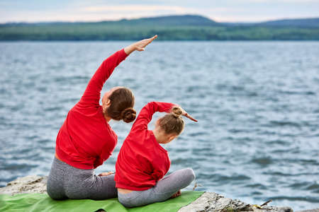 Young mother and little daughter doing yoga exercises on the stone near river.の写真素材