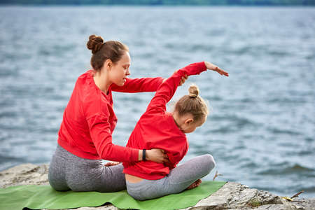 Rear view of young mother and daughter doing yoga exercises on the stone near the river. Beautiful mother and daughter in the sportwear and barefoot. Beautiful landscape like a background. Concept of the haelthy lifestyle and friendly family.の写真素材