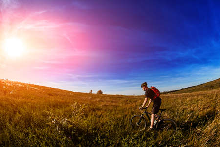 Young cyclist riding mountain bicycle through green meadow against beautiful sky.の写真素材
