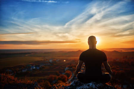 Rear view of the man meditating yoga in lotus pose on the rock at sunset. Horizontal photo....