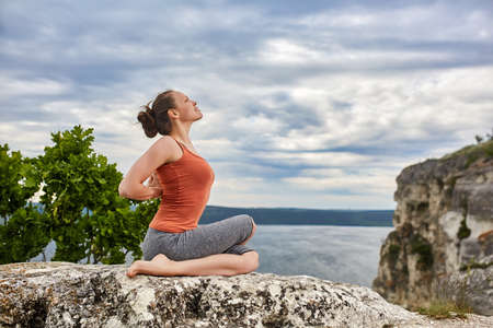 Portrait of the attractive woman is practicing yoga on the rock above river.