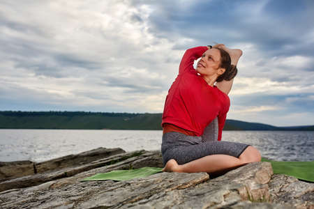 Happy young woman doing yoga exercise outdoors on the stone near river.