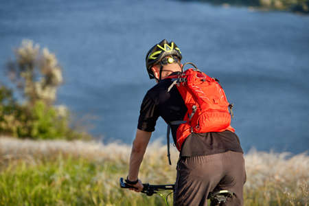 Young man riding mountain bike on the green meadow above the blue river in the countryside.の写真素材
