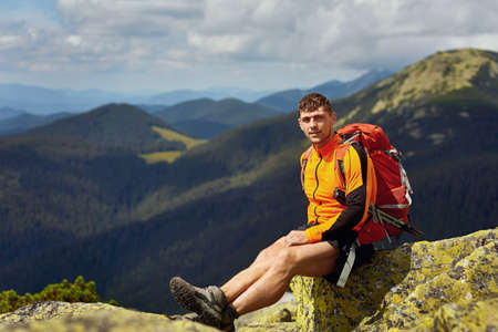 Young male hiker with backpack relaxing on top of a mountain during calm summer sunset