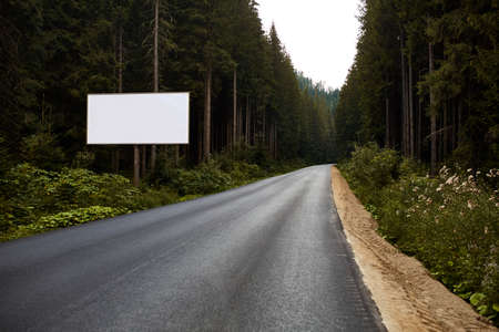 Blank billboard sign by empty highway through forest mountains landscape. Ad panel on road.の写真素材