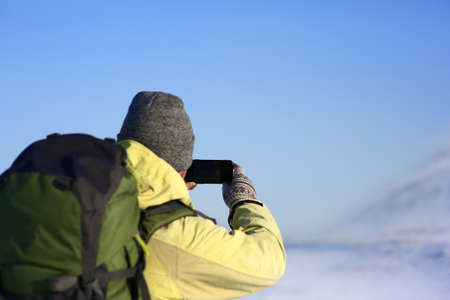 Back view of unrecognizable male in hat and cout with fur hood carrying stylish backpack taking picture of amazing winter landscape using copyspace mobile phone while walking alone on sunny day.の写真素材