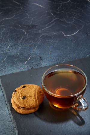 A transparent glass cup of black tea with chocolate chips cookies on a dark greyish marble background, vertical. Delicious dessert and refreshing teatime. Breakfast background.の写真素材