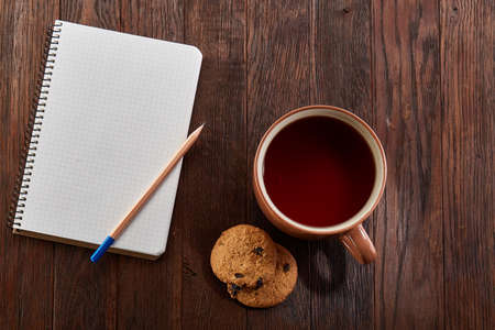 A white inside and brown outside porcelain cup of tea with tasty chocolate chips cookies, empty workbook and long yellow pencil with blue top on a dark wooden vintage background, top view.の写真素材