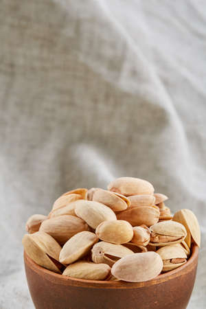 Composition of raw unpeeled pistachios in clay ceramic bowl on light grey cotton tablecloth or...